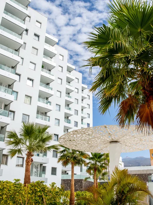 Facade of the white apartment complex rising above palm gardens, its balconies fitted with lace-like perforated panels.
