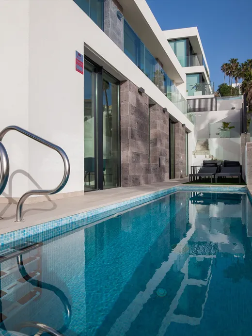 Narrow pool running beside the white stepped villas, with a glass balustrade and hillside beyond.