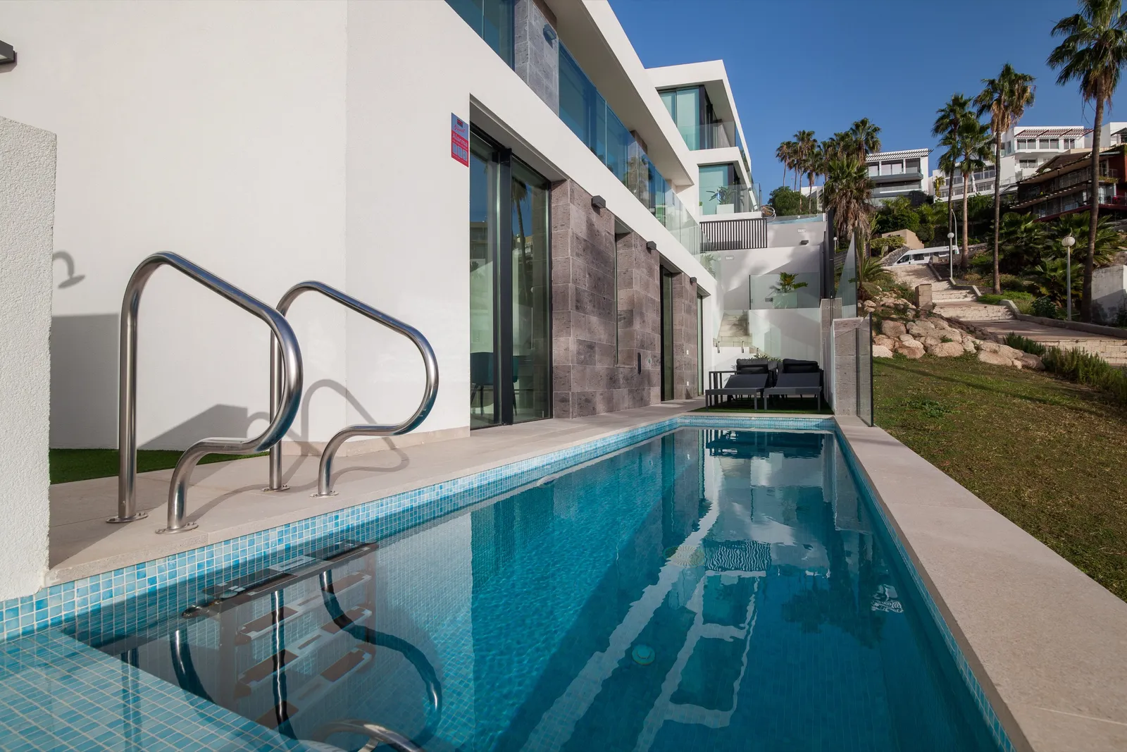 Narrow pool running beside the white stepped villas, with a glass balustrade and hillside beyond.