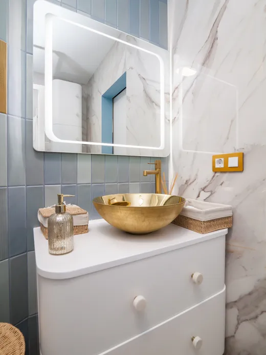 Bathroom with a brass vessel sink on a white vanity, marble and pale blue tiles and an LED mirror.