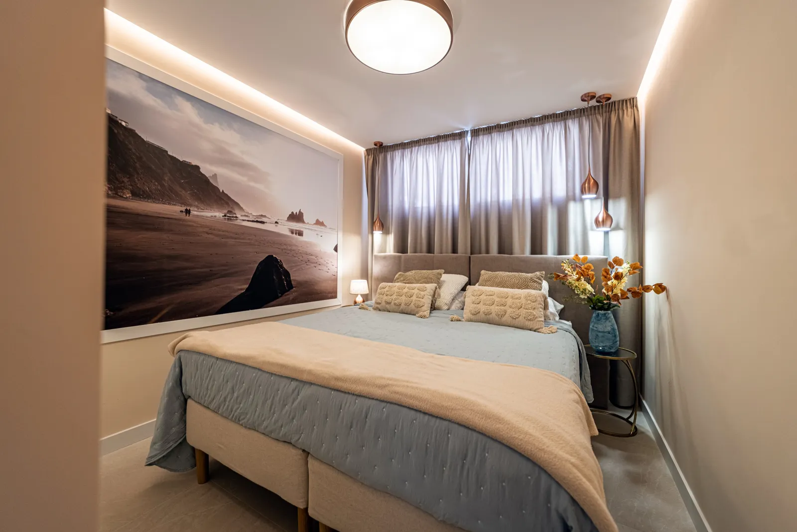 Bedroom with a black-sand beach photo mural above the bed, copper pendant lights and warm neutral bedding.