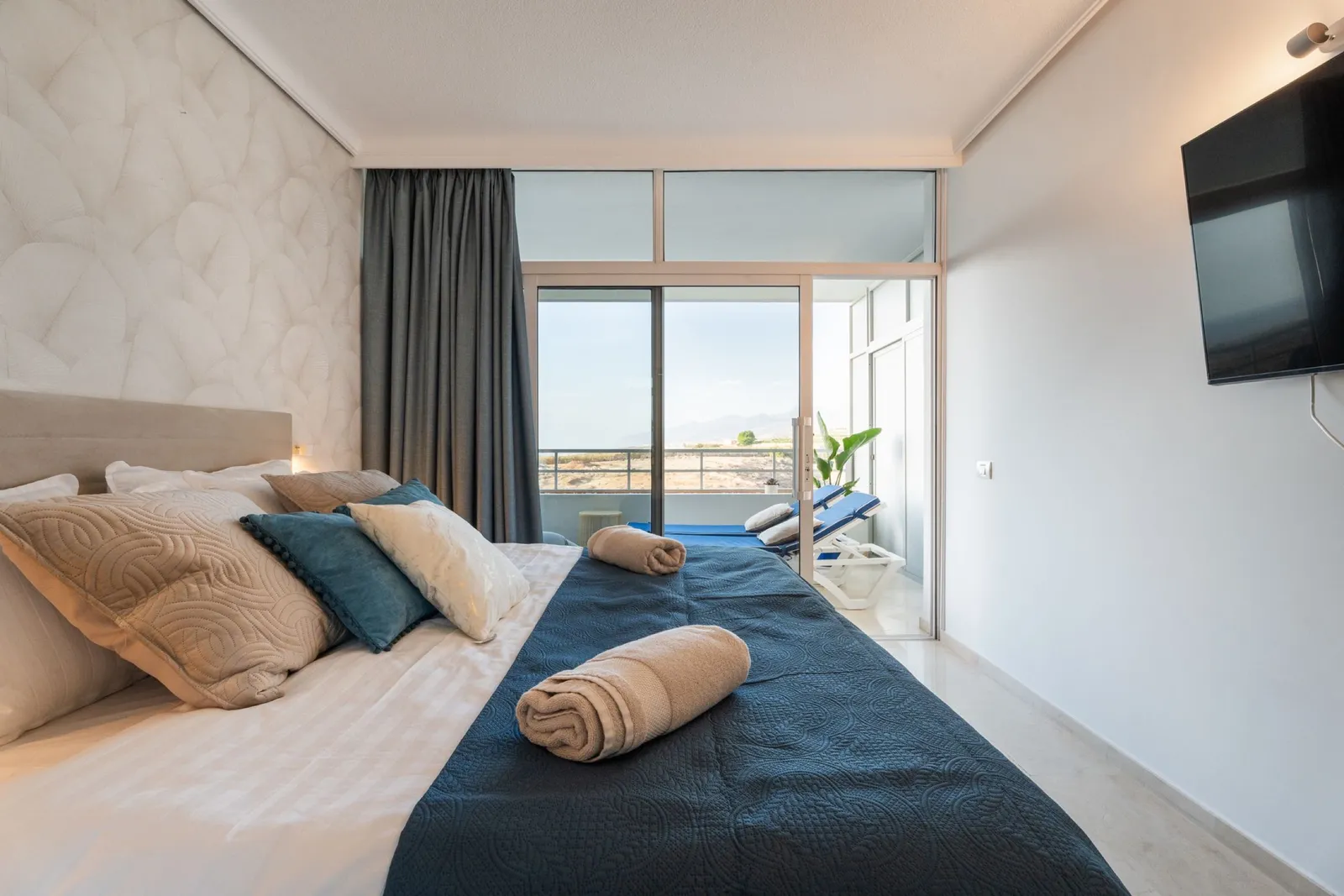 Dormitorio con un balcón con vistas al mar, ropa de cama en azul marino y beige y un televisor de pared.
