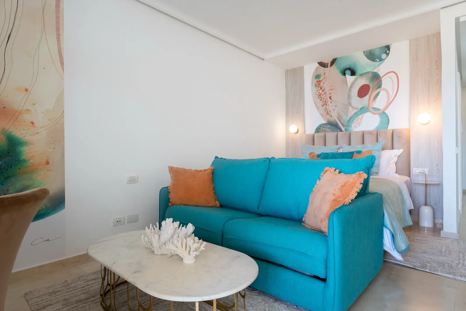 Rincón de comedor con sillas de terciopelo color camel y un mural fluido en coral y verde, junto a una ventana con vistas al mar.