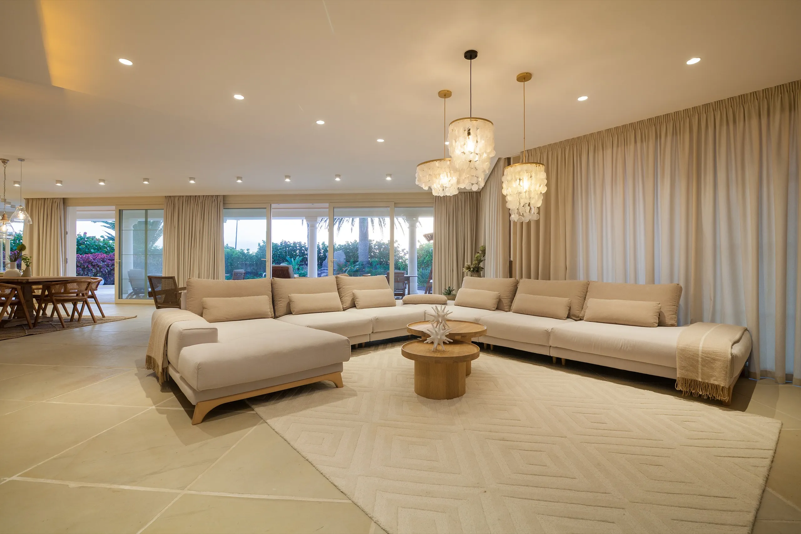 Amplio salón en tonos neutros con sofás color crema, lámparas de nácar y cortinas de suelo a techo abiertas a un jardín.