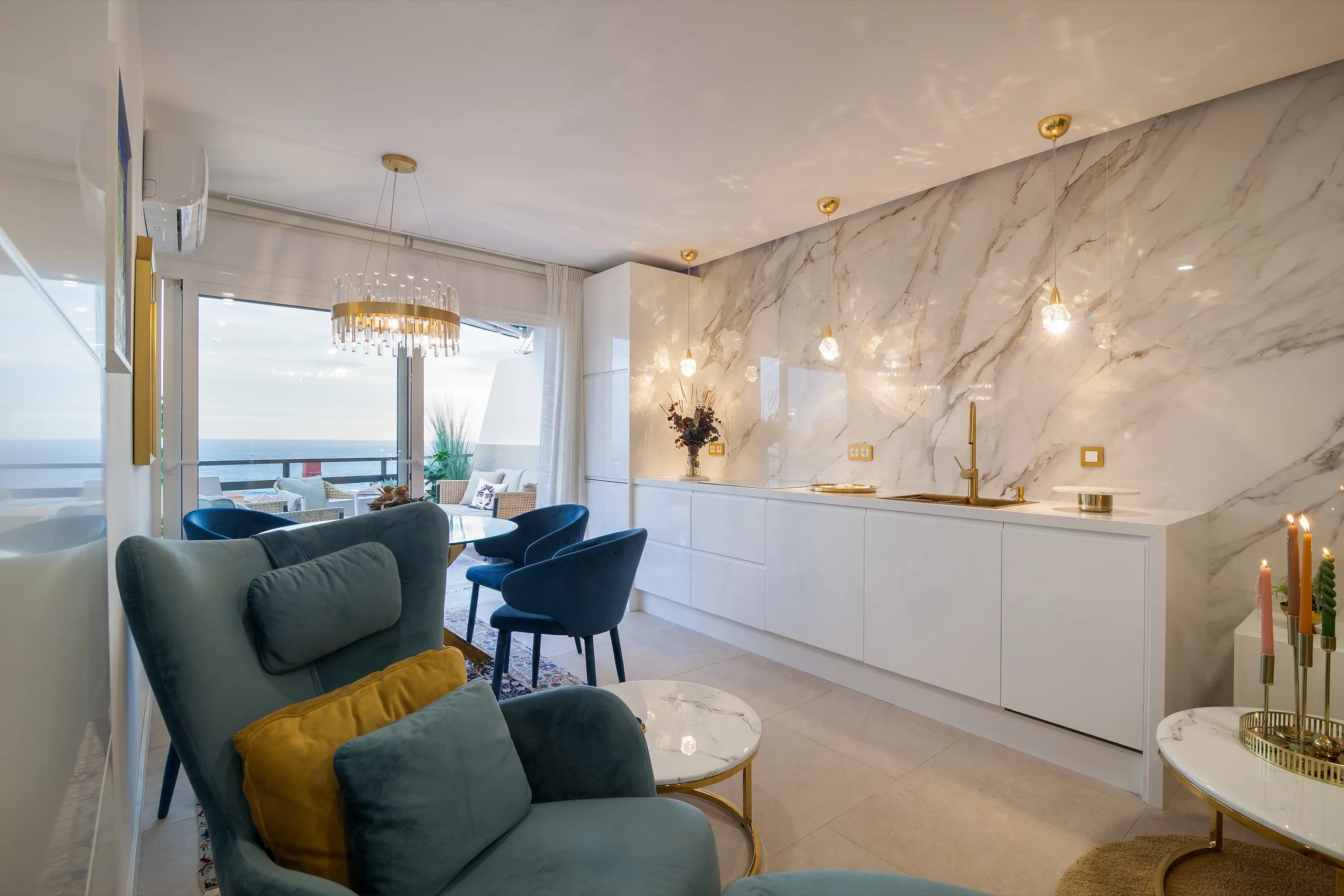 Zona de salón con una pared de mármol, griferías de latón, sillones turquesa y una vista al mar.