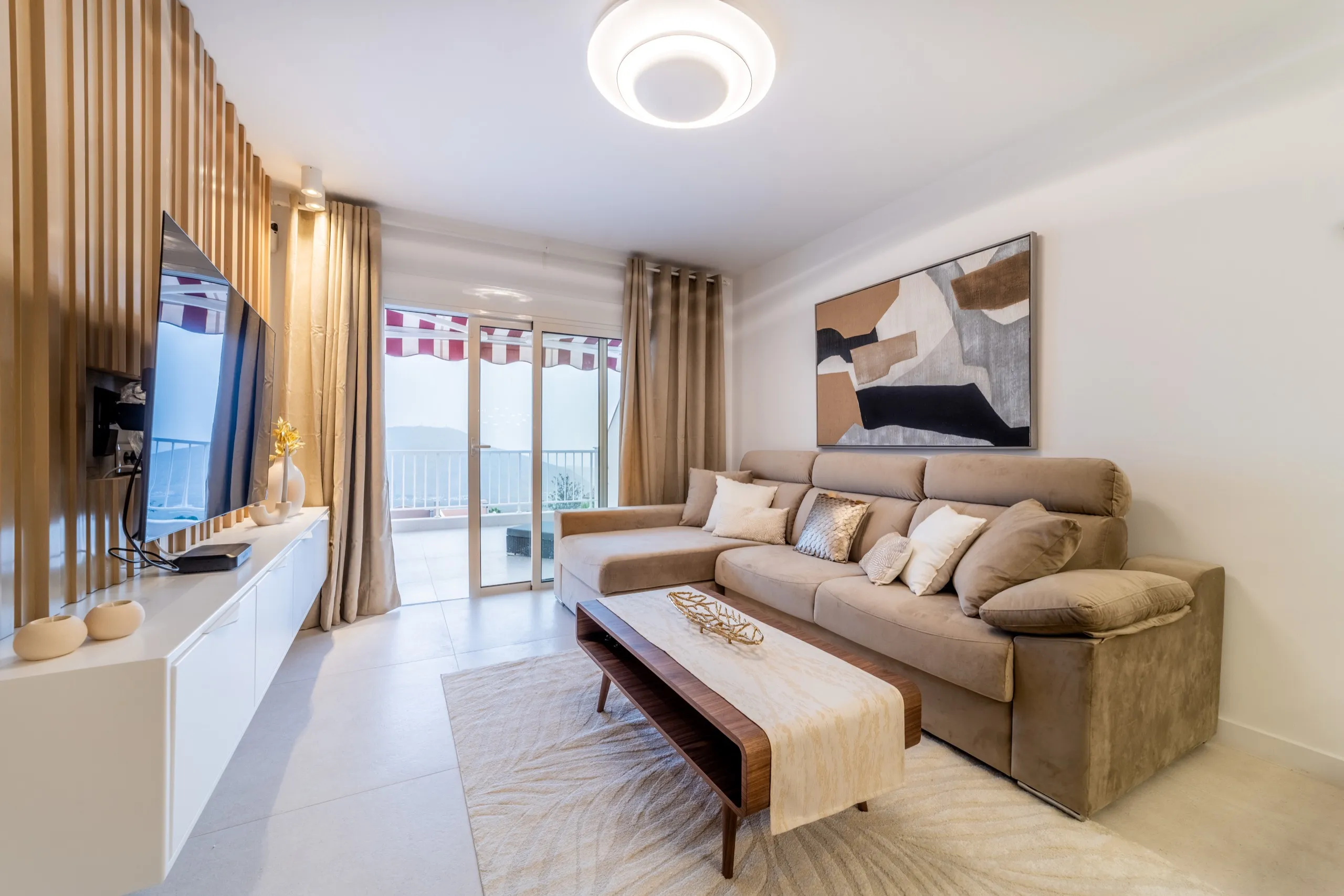 Salón en beis claro y roble con un sofá suave, arte abstracto en marrones y negros apagados y un balcón con vistas al mar.