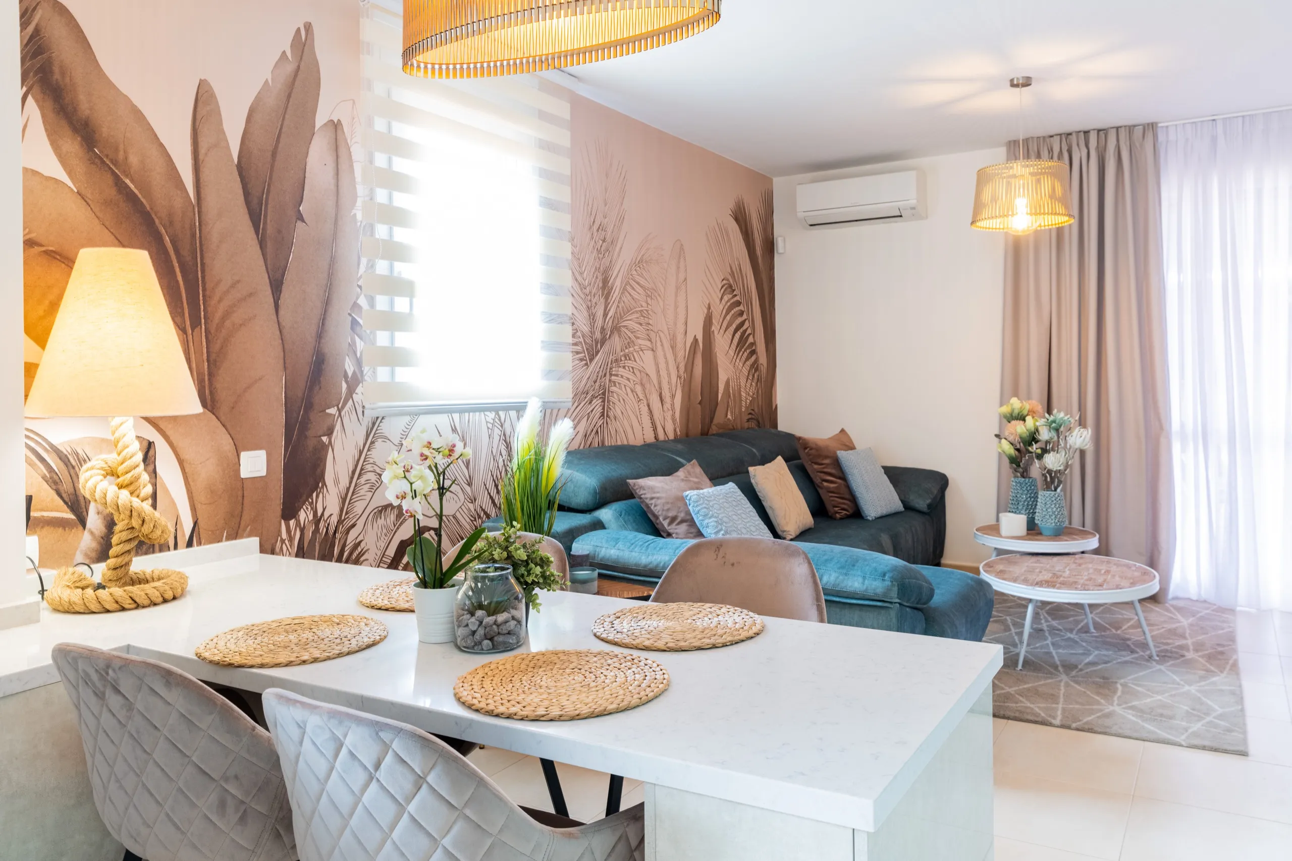 Salón-comedor abierto con un mural tropical de palmeras en marrón suave, un sofá azul y una isla de cocina blanca.