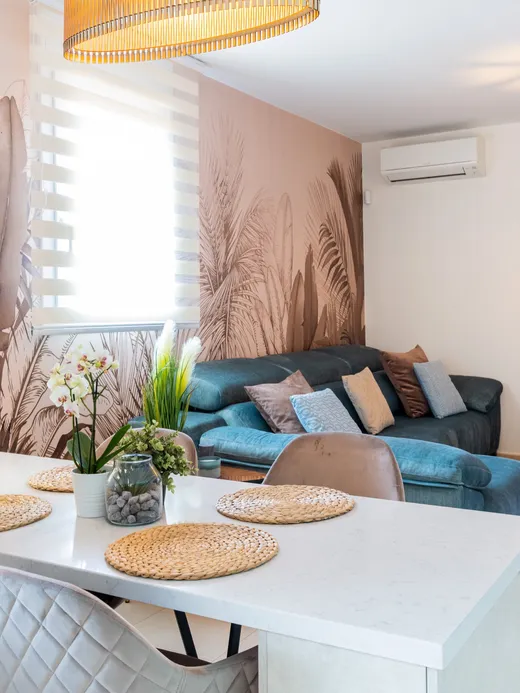 Open living and dining space with a soft brown tropical-palm mural, a blue sofa and a white kitchen island.
