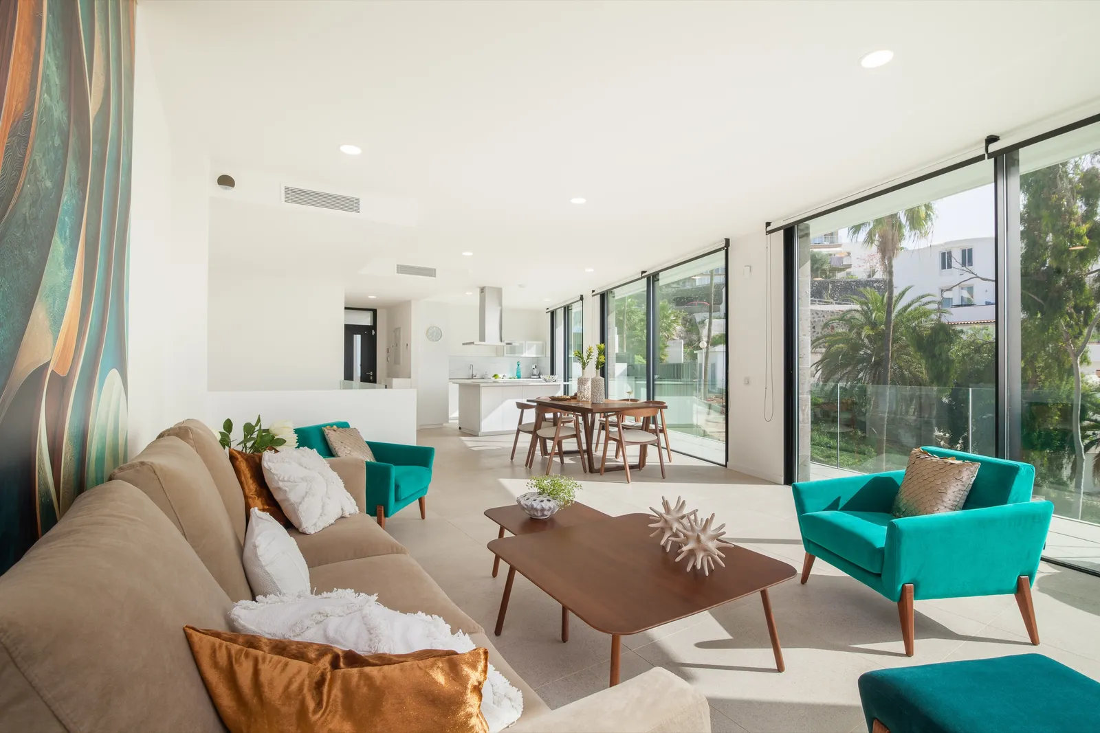 Open-plan living room with a linen sofa, turquoise accent chairs, camel leather cushions and a timber dining table, opening through full-height glazing to the garden.