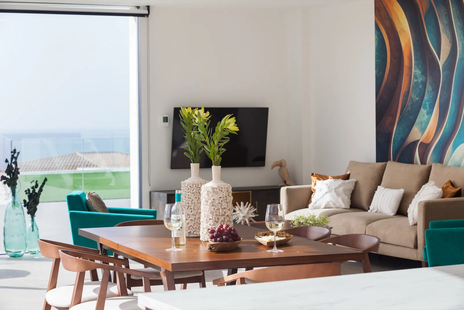Living and dining area with turquoise chairs and a fluid-art wall in blues and coppers, framed by floor-to-ceiling windows onto the terrace.